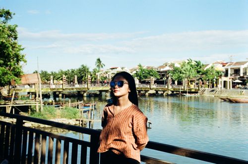 Free An Asian woman enjoying a sunny day by a riverside bridge, wearing sunglasses. Stock Photo