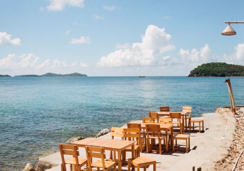 Free Rustic wooden tables and chairs overlook a serene ocean view, perfect for a summer vacation. Stock Photo