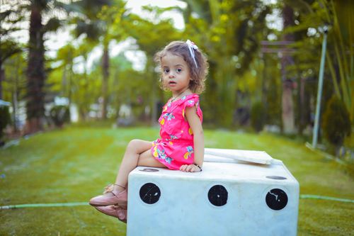 Free Cute toddler in a bright dress sitting outdoors on a white box surrounded by lush greenery in a park. Stock Photo