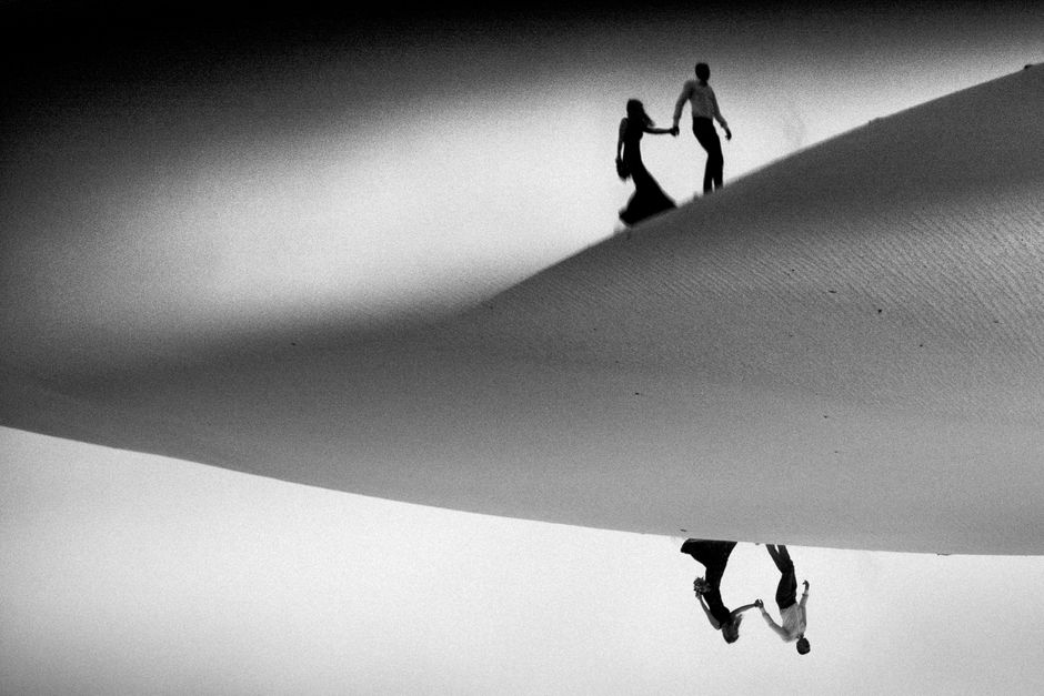 A creative black and white photo of two people walking and holding hands in a desert with a reflection.