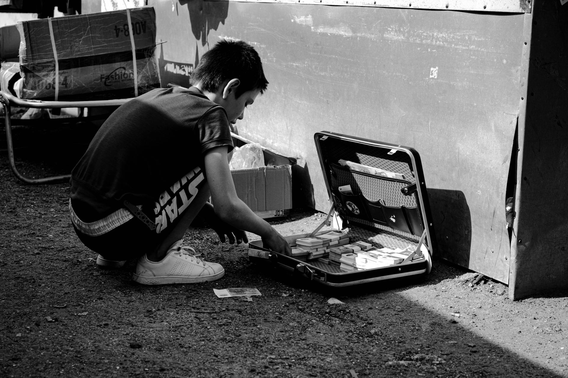 Black and white photo of a child examining money in a briefcase on a street.