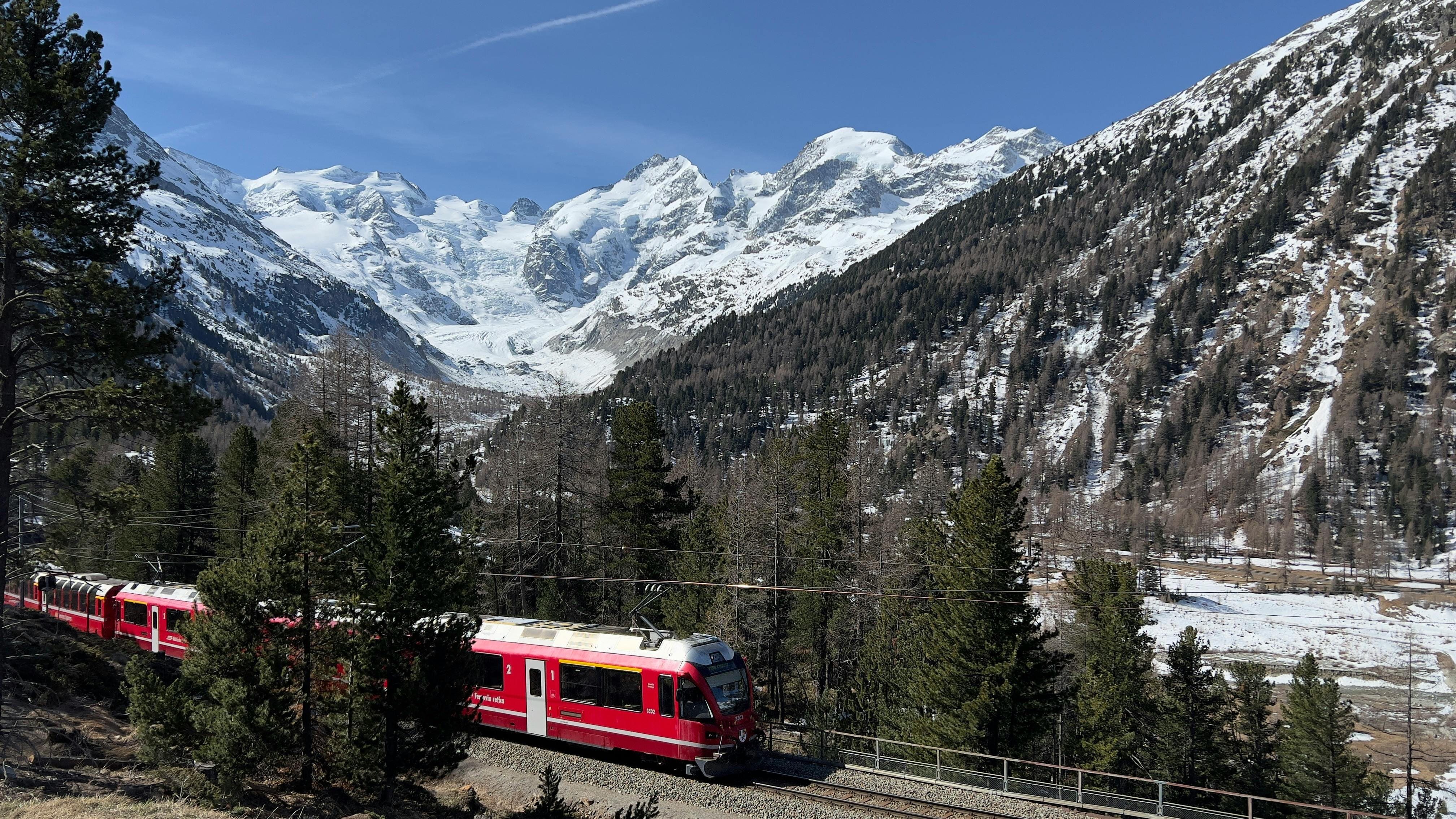 Red train travels through a snowy mountain landscape with pine trees and peaks in the background.