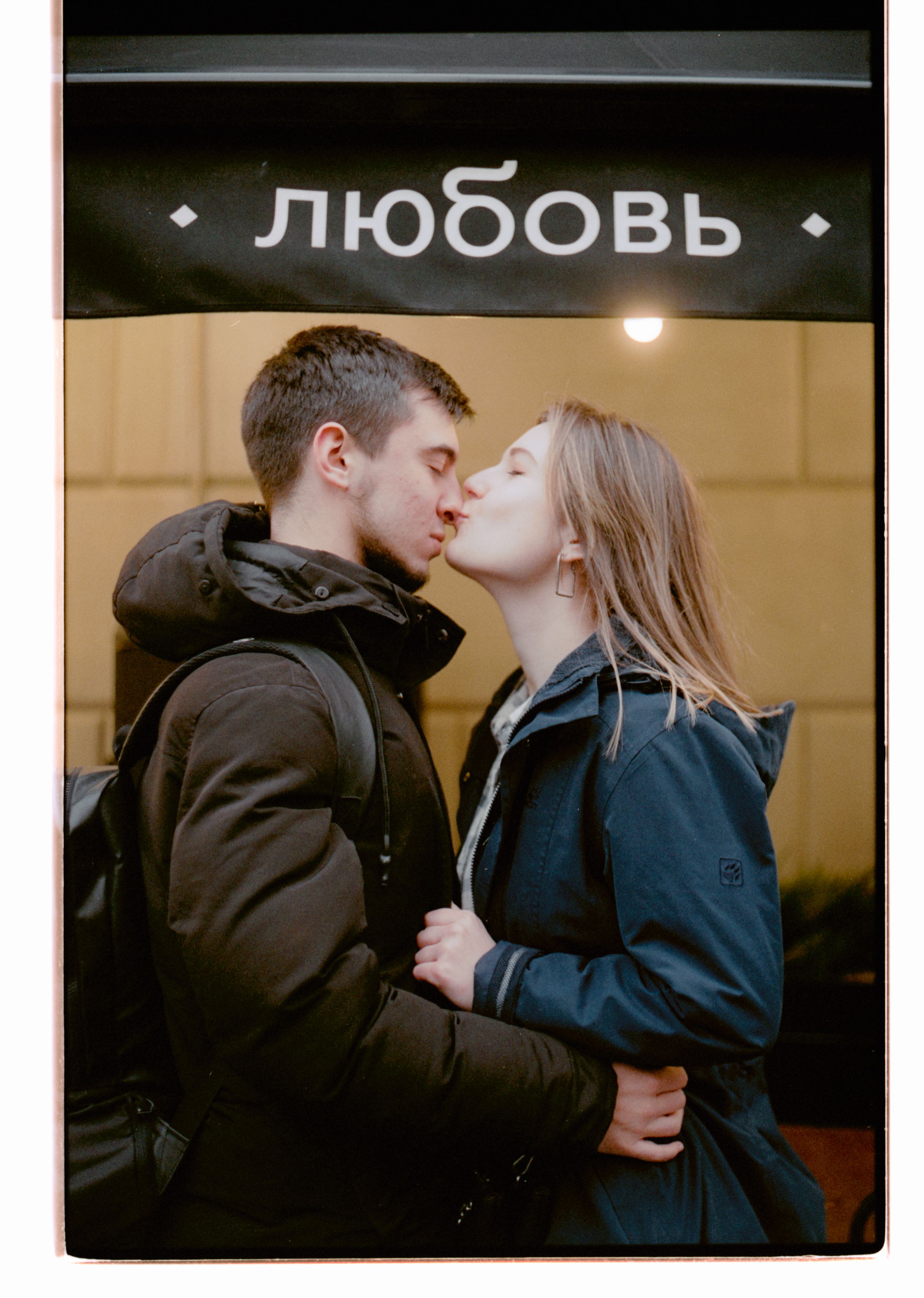 A young couple stands kissing under a sign with Cyrillic text, embracing warmly.