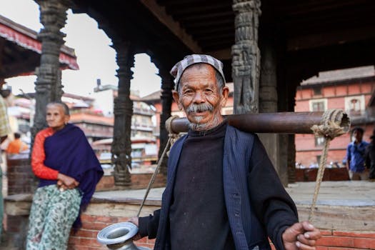 An elderly man smiles while carrying a traditional yoke in a vibrant outdoor market scene.