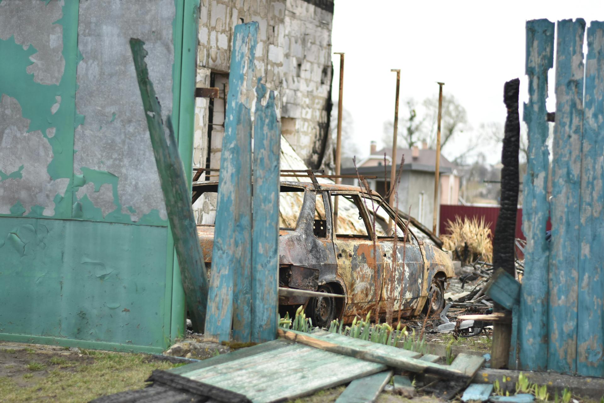 A rusted, abandoned car amidst a decaying urban environment with damaged fences and peeling paint.