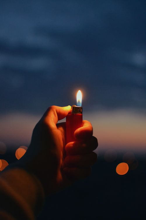 Free A person holding a lighter with a flame, against a twilight sky with bokeh lights. Stock Photo