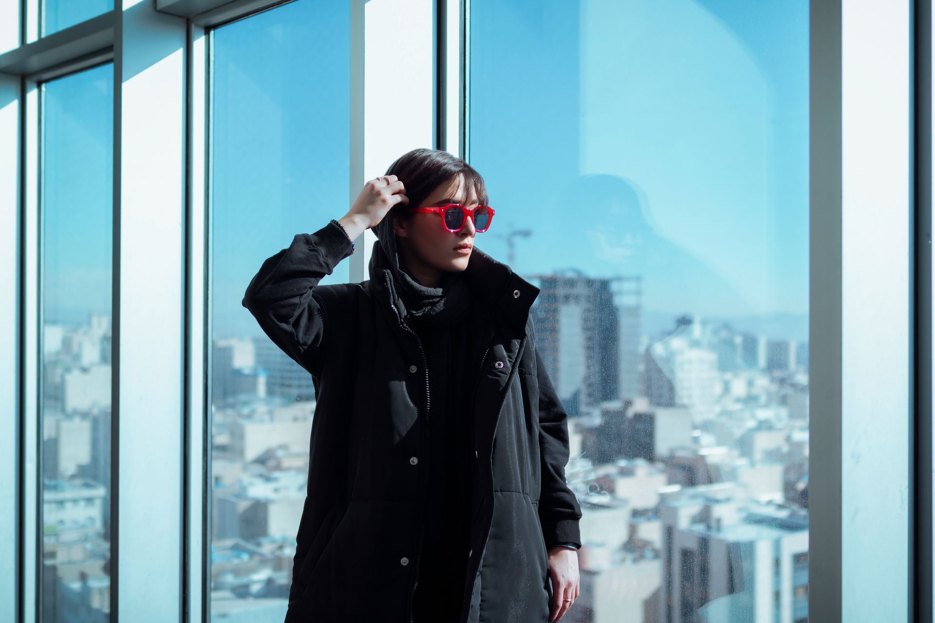 Woman wearing red sunglasses gazes over Tehran skyline from window, capturing modern urban fashion.