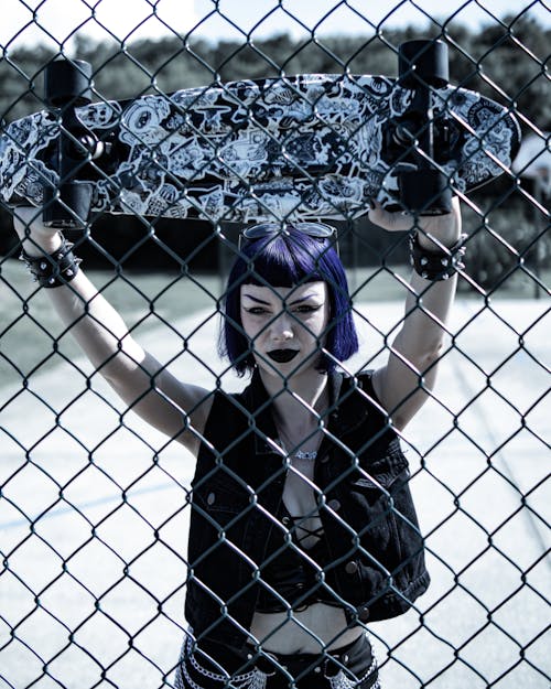 Woman Holding Up a Skateboard Beside a Chain Link Fence