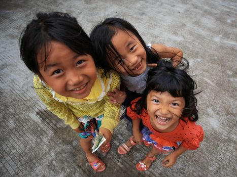 Three young girls smiling joyfully while standing outdoors on a pavement, captured from above.