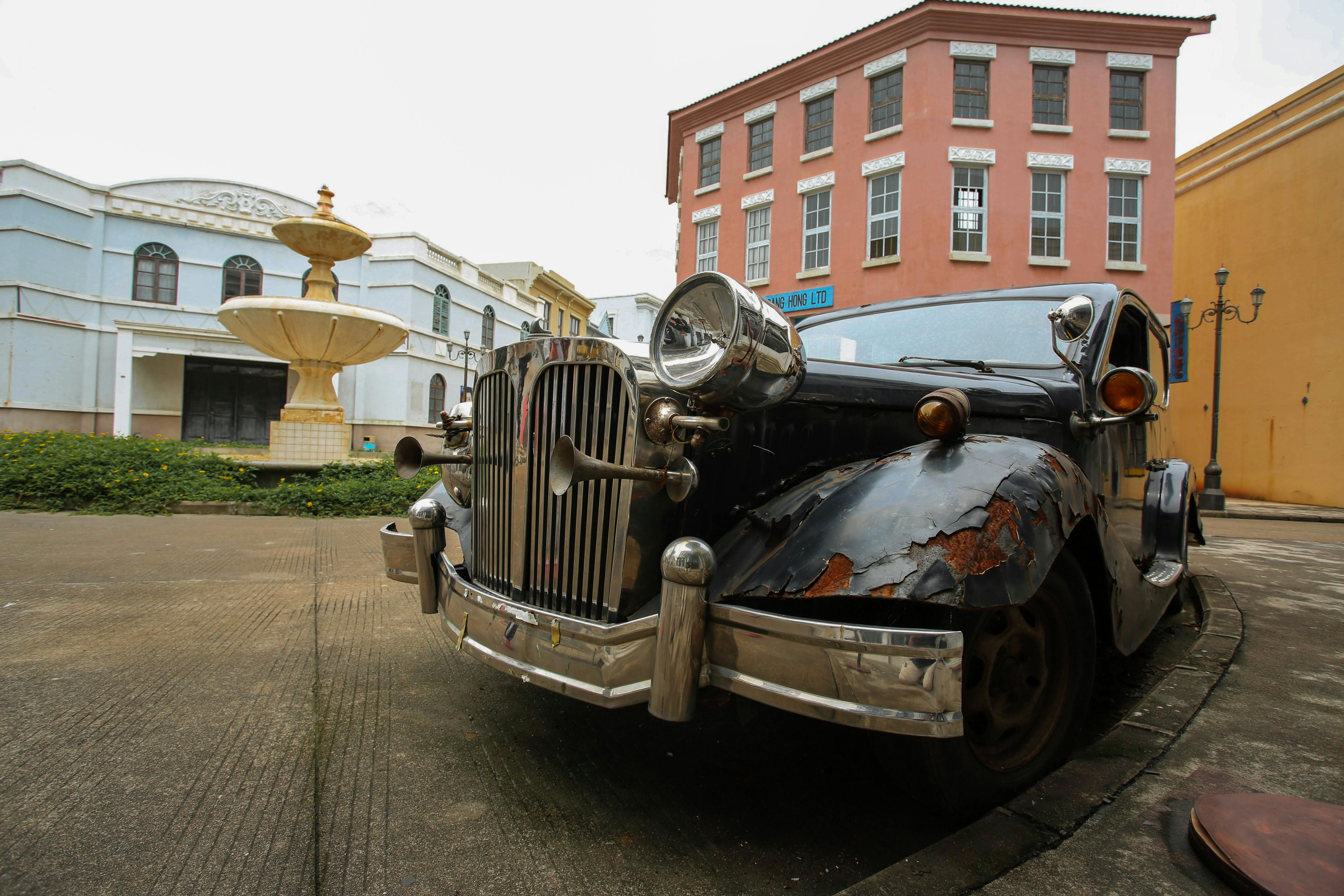 A classic rusty car parked near historical buildings and a fountain.