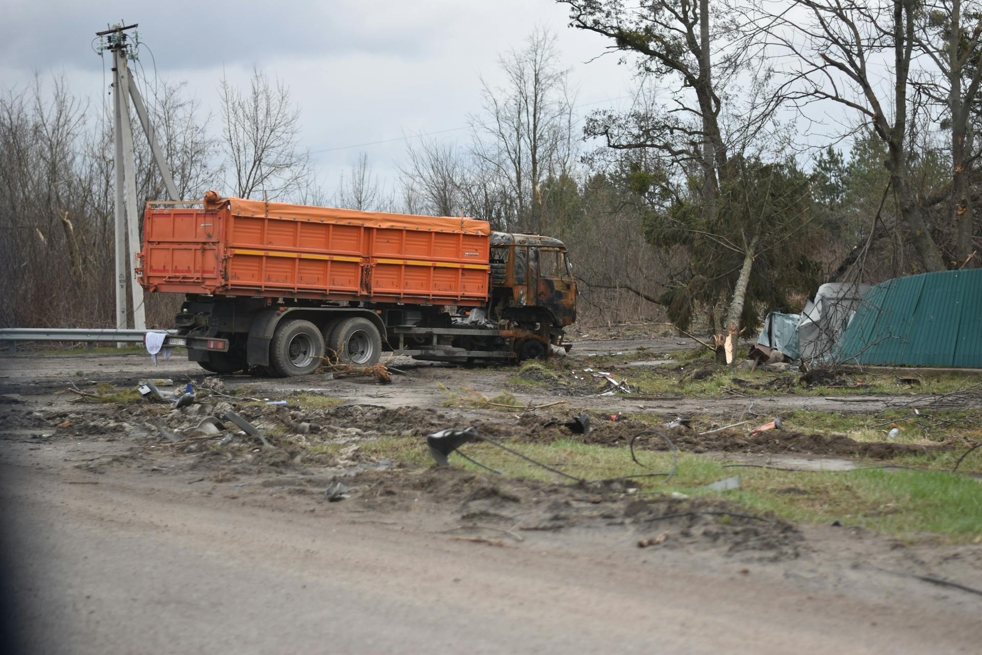 An abandoned, damaged truck on a muddy road surrounded by debris and barren trees.