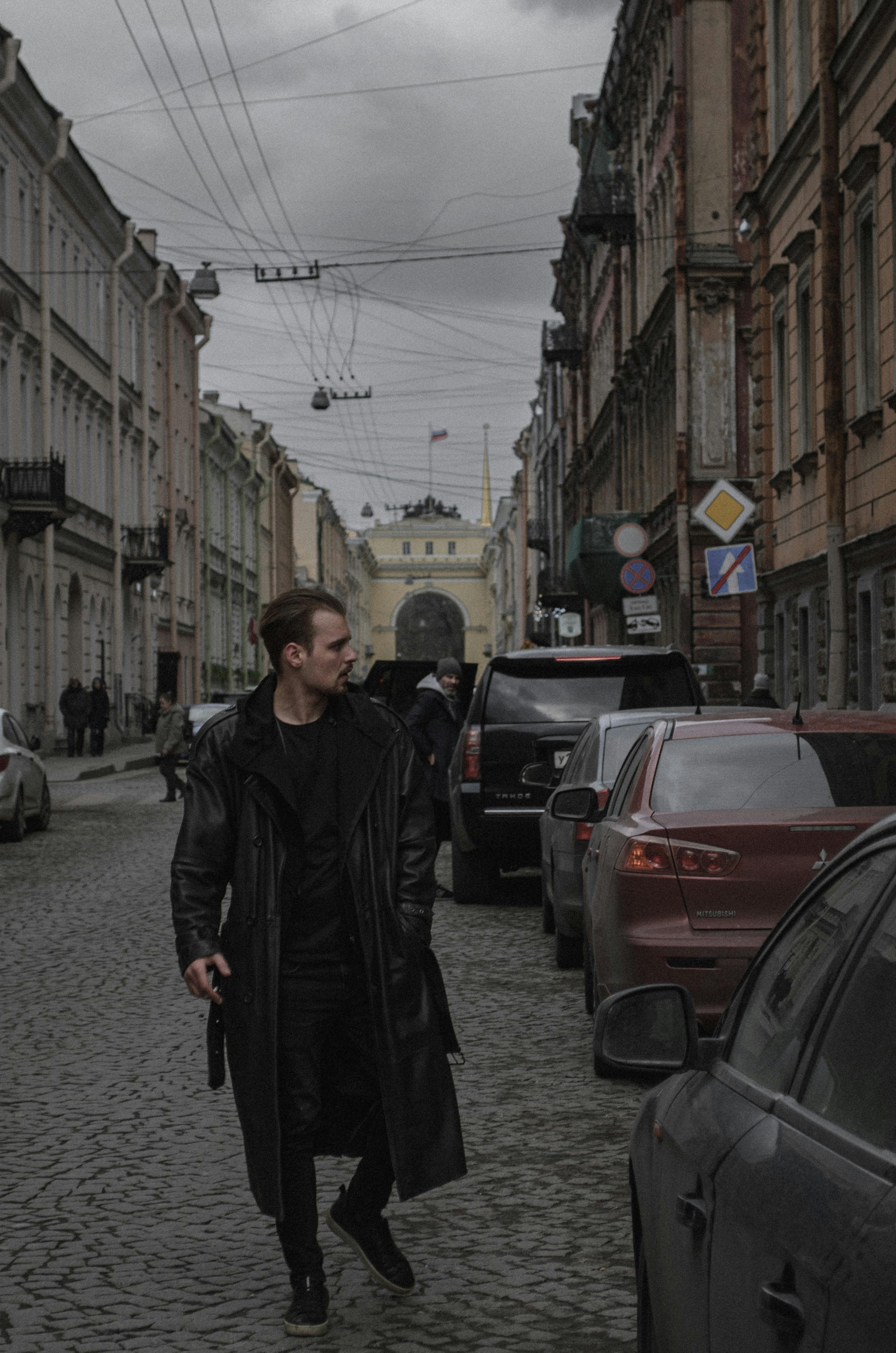 A man in a long black coat walking down a cobblestone street in a historical city setting.