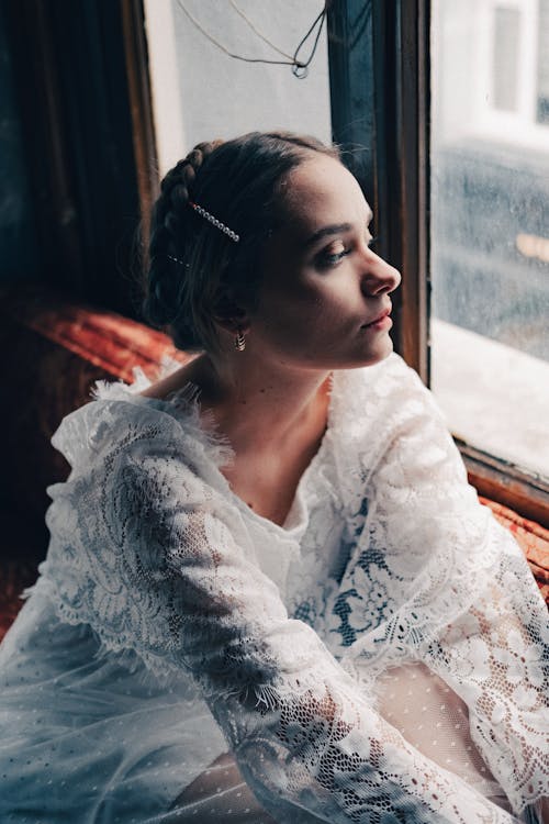 Free Woman in White Lace Dress Sitting and Looking Through the Window Stock Photo