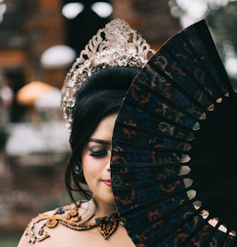 A woman in traditional attire elegantly holding a fan, showcasing intricate jewelry and headpiece.