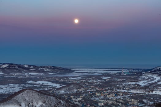 A serene winter cityscape with mountains and a full moon in twilight.
