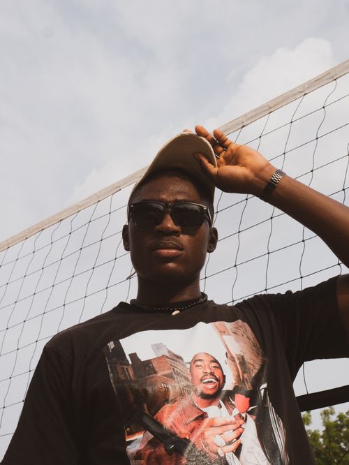 Free Portrait of a stylish Nigerian man wearing sunglasses and a cap, standing outdoors by a net. Stock Photo