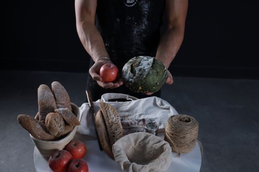 A man displays artisanal bread and a red apple with rustic food items on a table.
