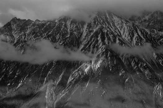 Black and white image of snow-covered mountains shrouded in clouds and fog under an overcast sky.