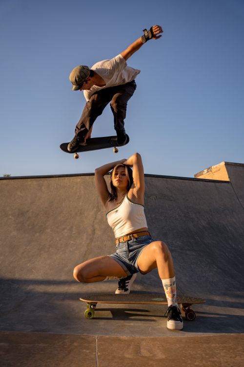Free A skateboarder performs an aerial trick over a woman posing at a skatepark under clear skies. Stock Photo