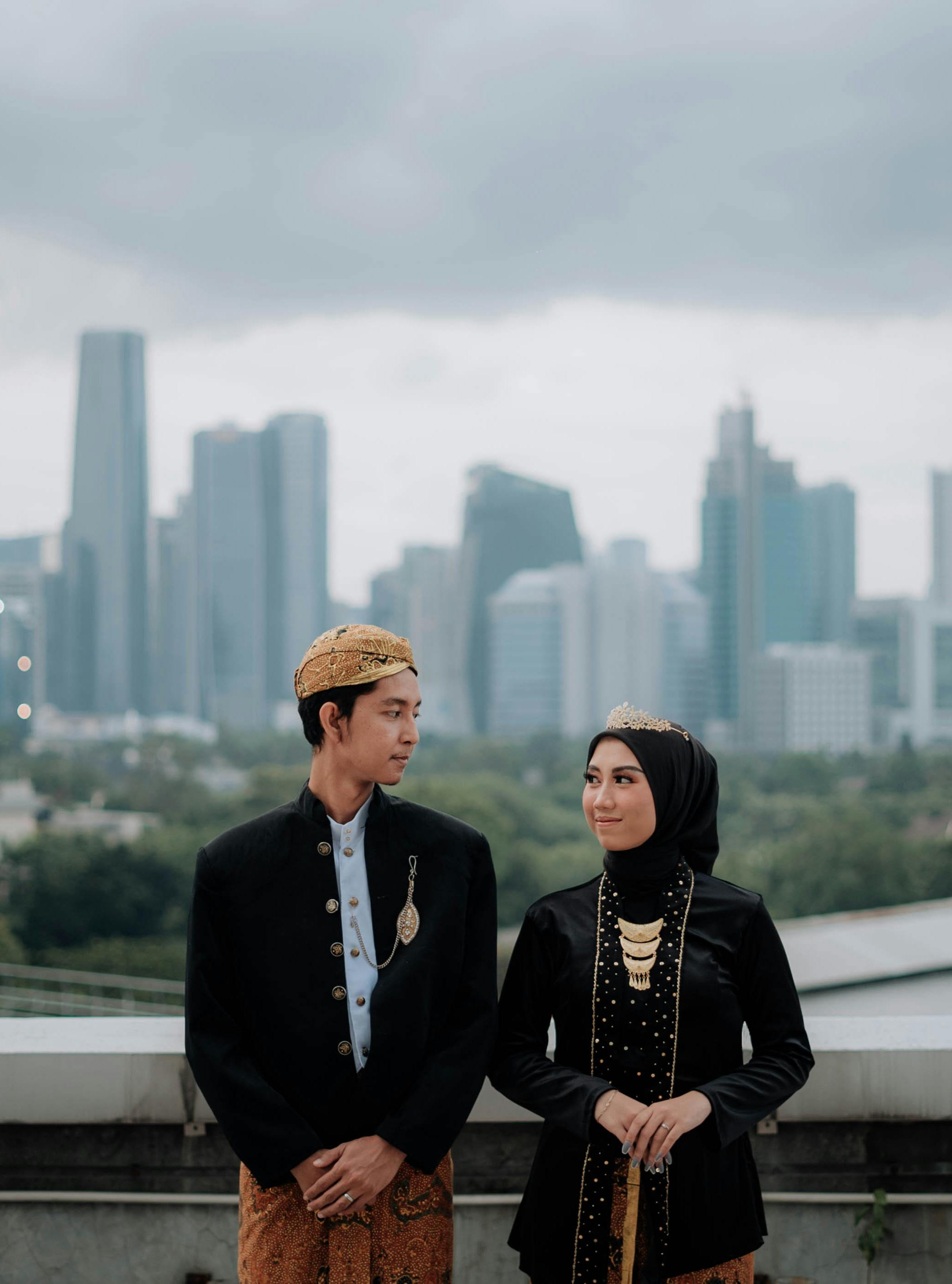 A couple in traditional Indonesian clothing posing with a modern cityscape in the background.