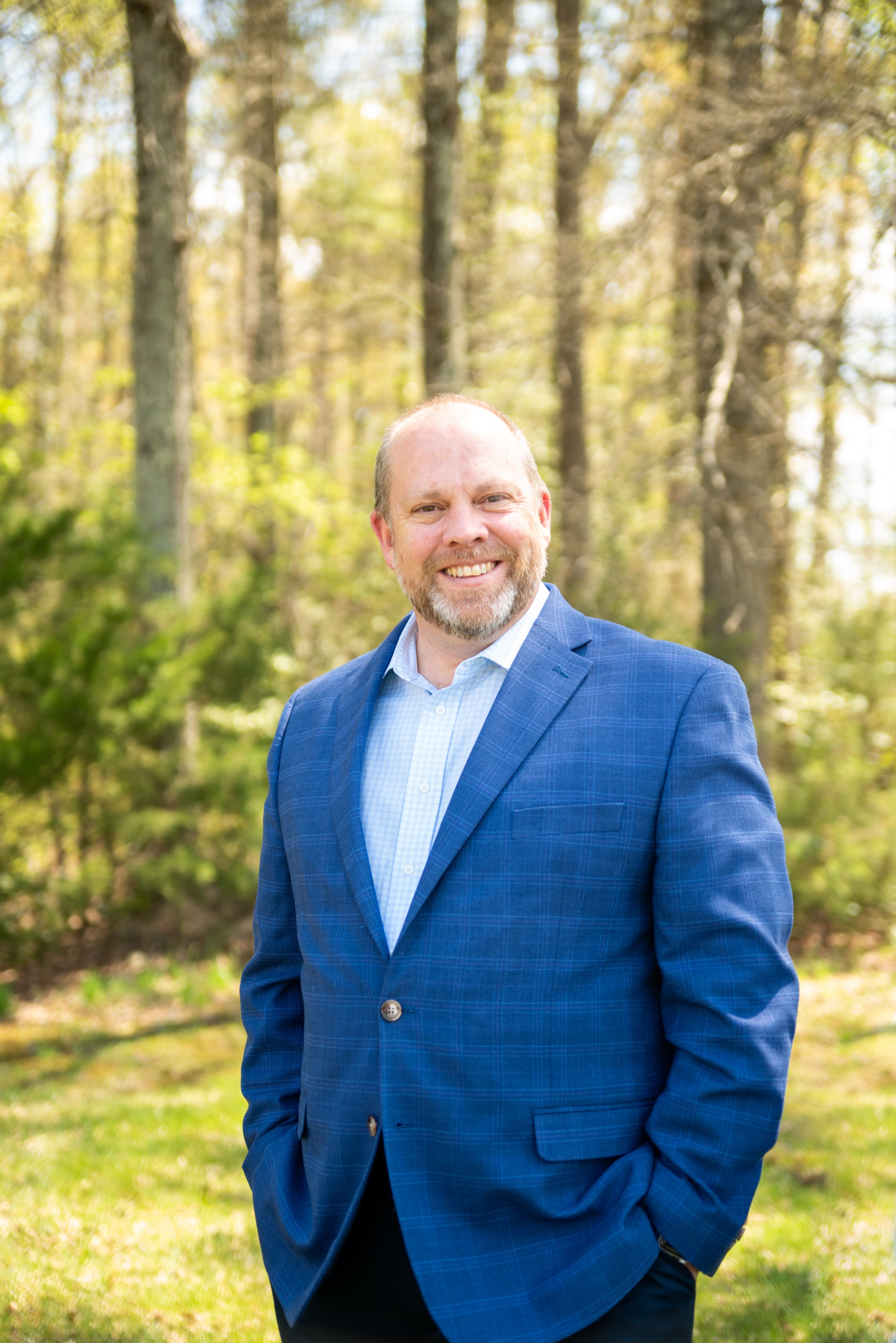 Smiling man in blue suit jacket standing outdoors in a sunny wooded area.