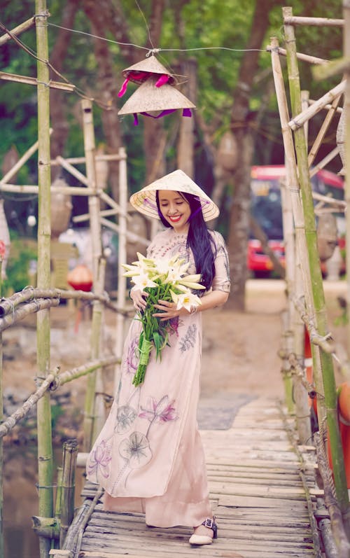 Woman in a Floral Dress Holding a Bouquet of White Flowers while on a Wooden Bridge