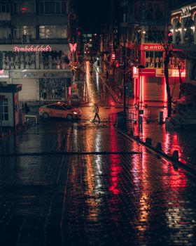 Dramatic night street scene with red neon lights and reflections on wet pavement, featuring a taxi and pedestrian.