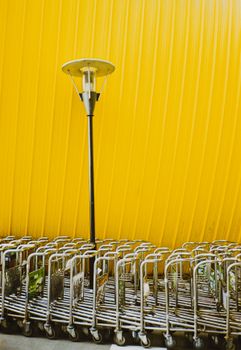 Industrial metal luggage carts lined up against a vibrant yellow wall under a lamppost.