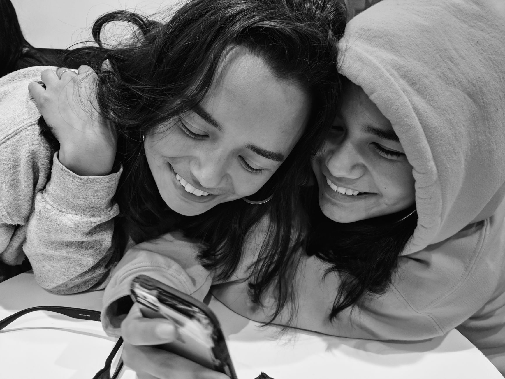 Two teenage girls laughing and smiling while looking at a smartphone in a black and white photo.