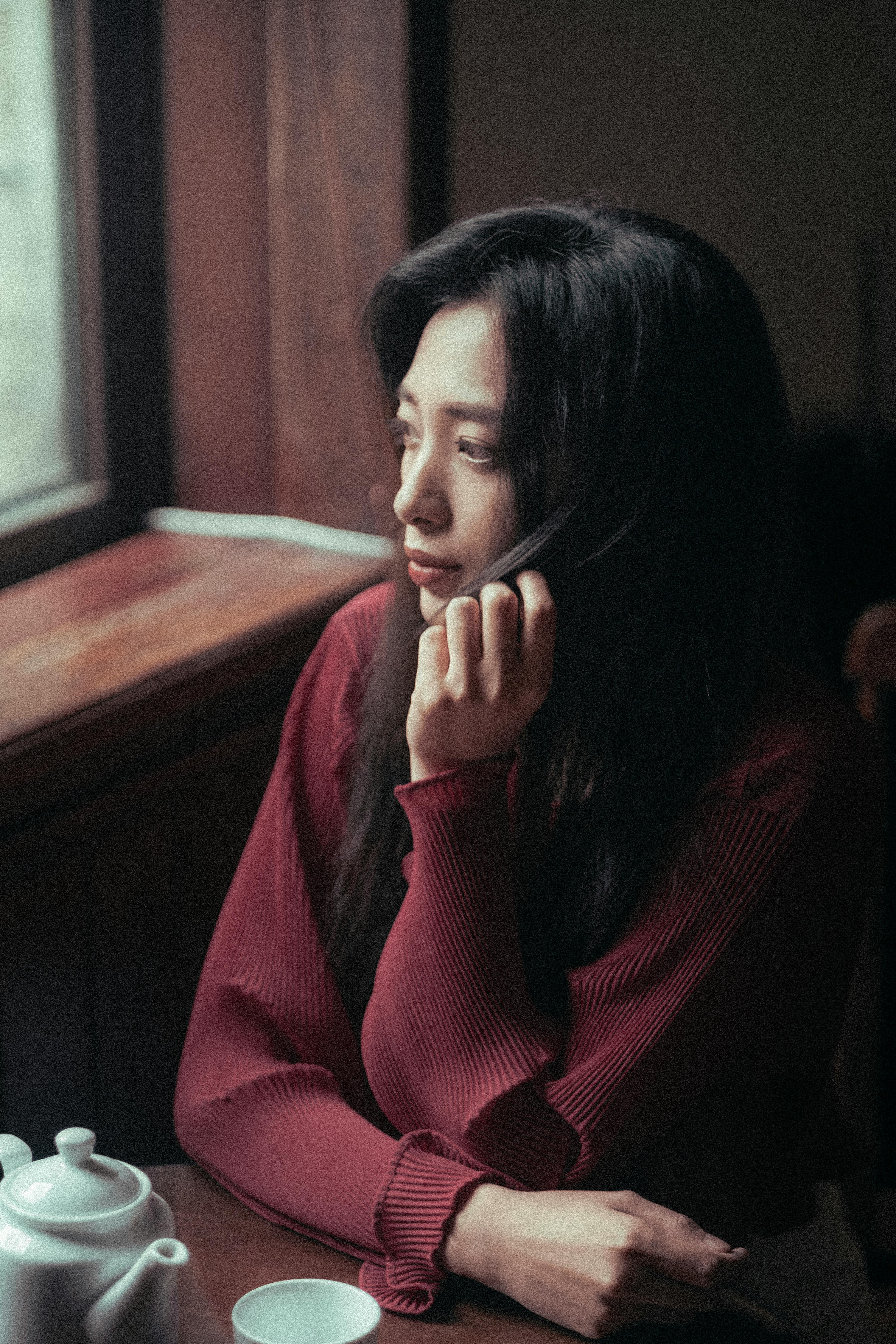Thoughtful Asian woman in maroon sweater gazing out window, evokes serene introspection.