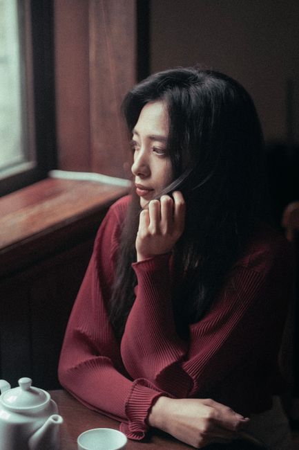Thoughtful Asian woman in maroon sweater gazing out window, evokes serene introspection.