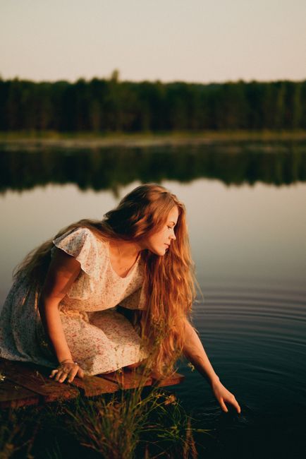 A serene scene of a young woman with long hair reaching out to touch a tranquil lake during sunset.