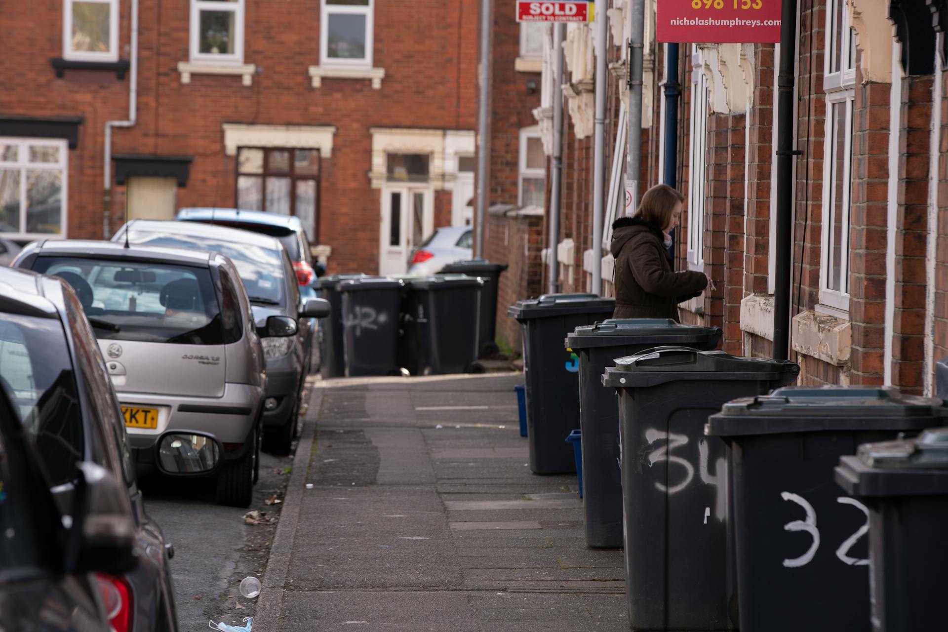 A street scene in a residential area with garbage bins and parked cars along the sidewalk.