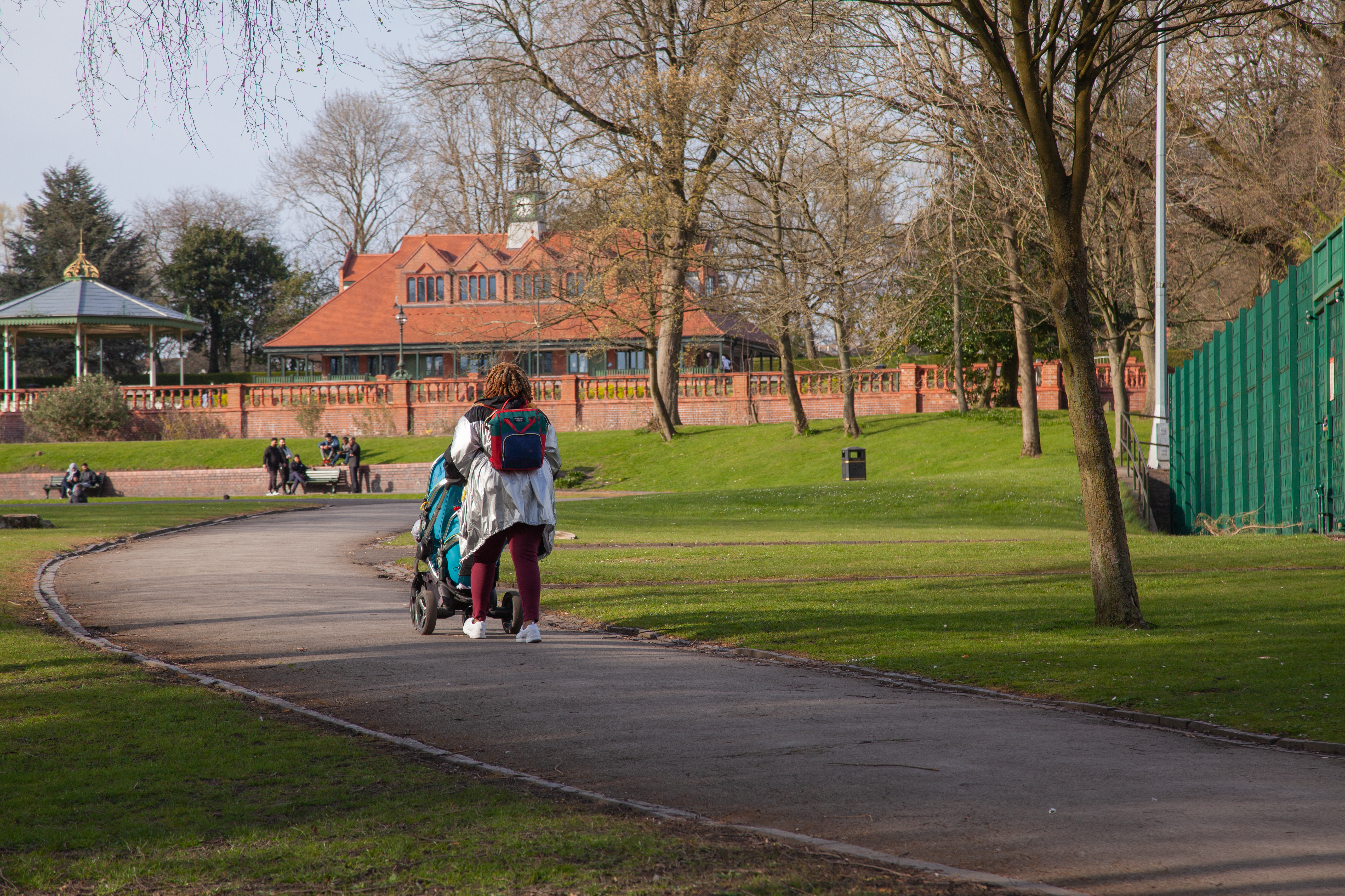 A woman pushes a stroller along a park pathway, surrounded by greenery and trees.
