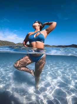 A woman in blue swimwear poses confidently in clear tropical waters under a bright blue sky.