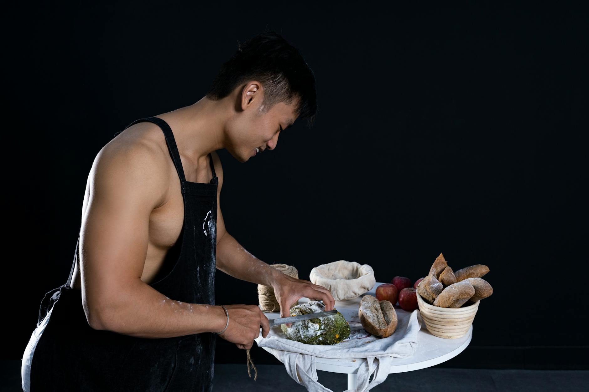 A muscular man in an apron slices freshly baked bread in a studio setting, exuding a fit lifestyle.