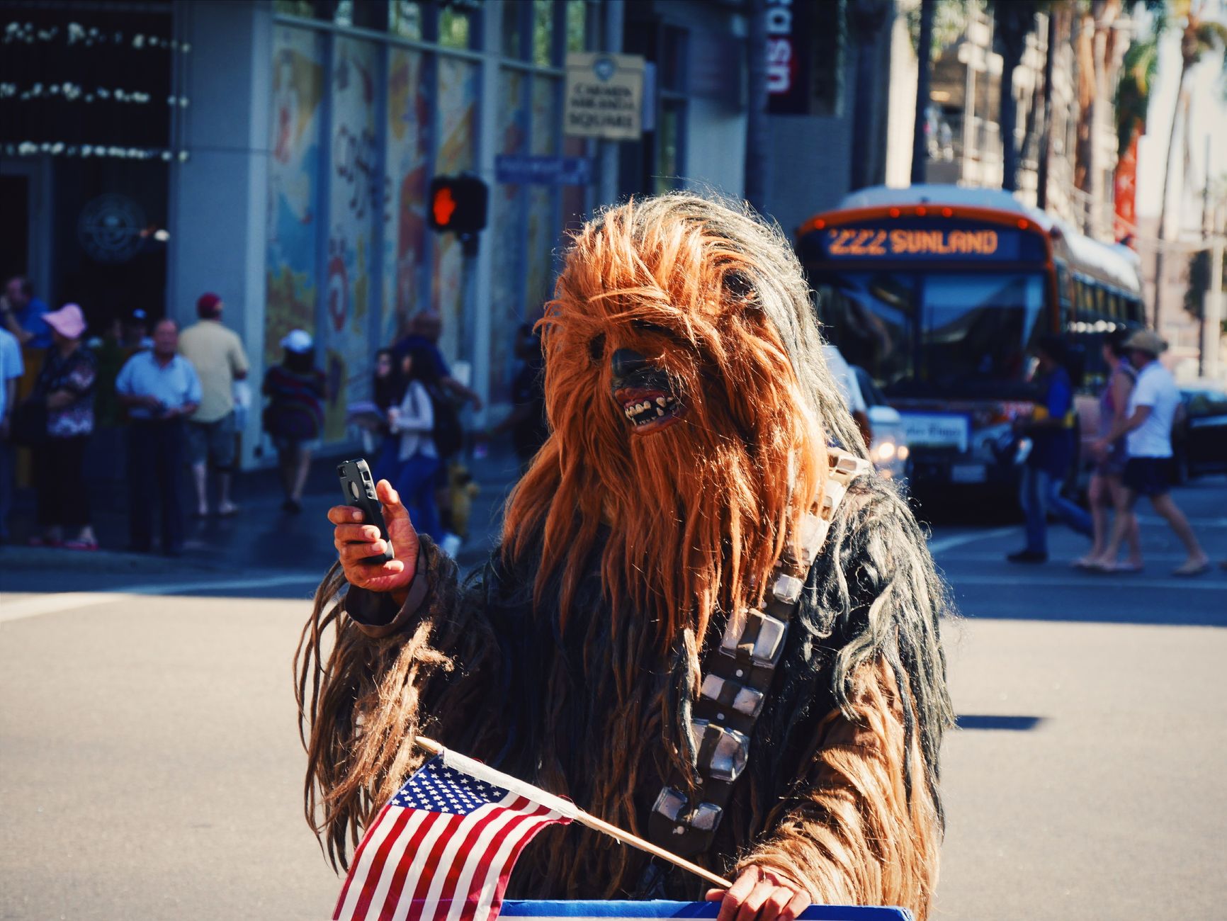 Chewbacca character cosplayer holding a phone and flag on a bustling city street.
