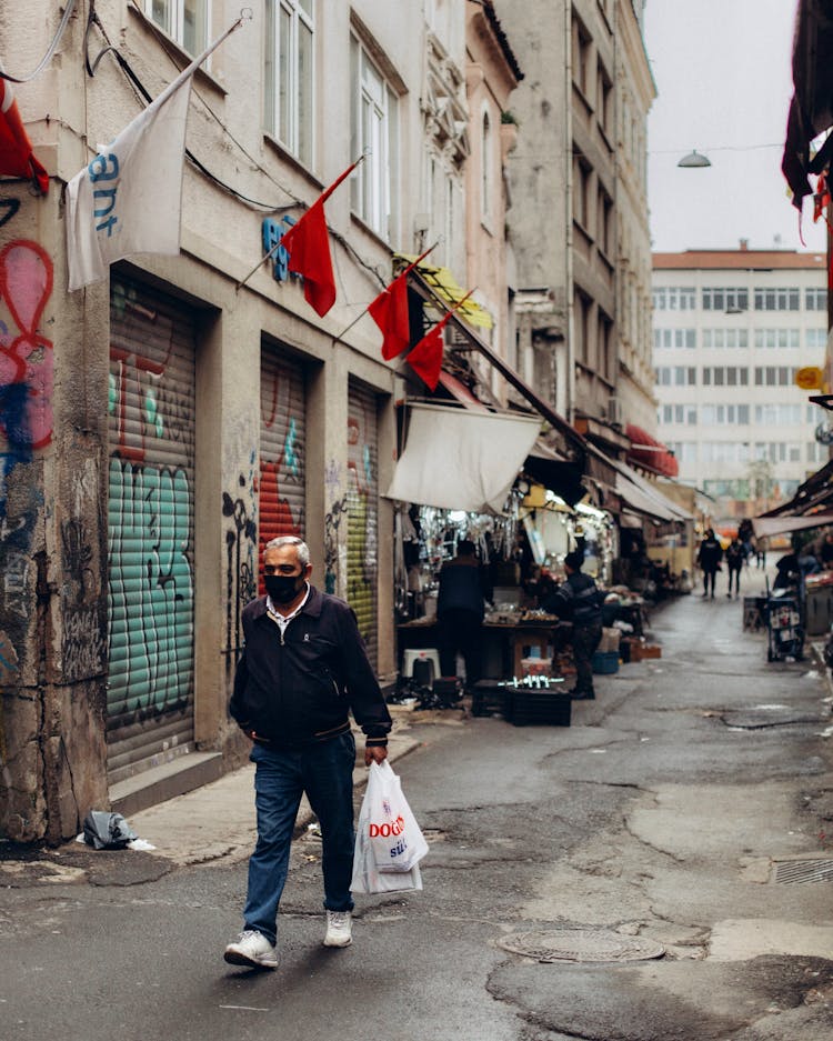 Man In Face Mask Walking City Street With Flags On Buildings