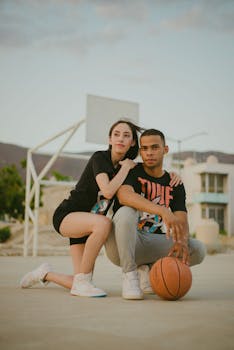 A young couple in casual attire sitting on an outdoor basketball court, showcasing sporty fashion.