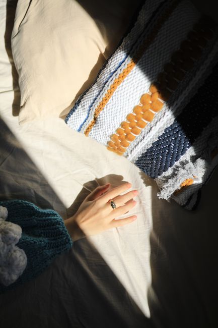 A hand wearing a ring rests on a sunlit bed with a textured pillow.