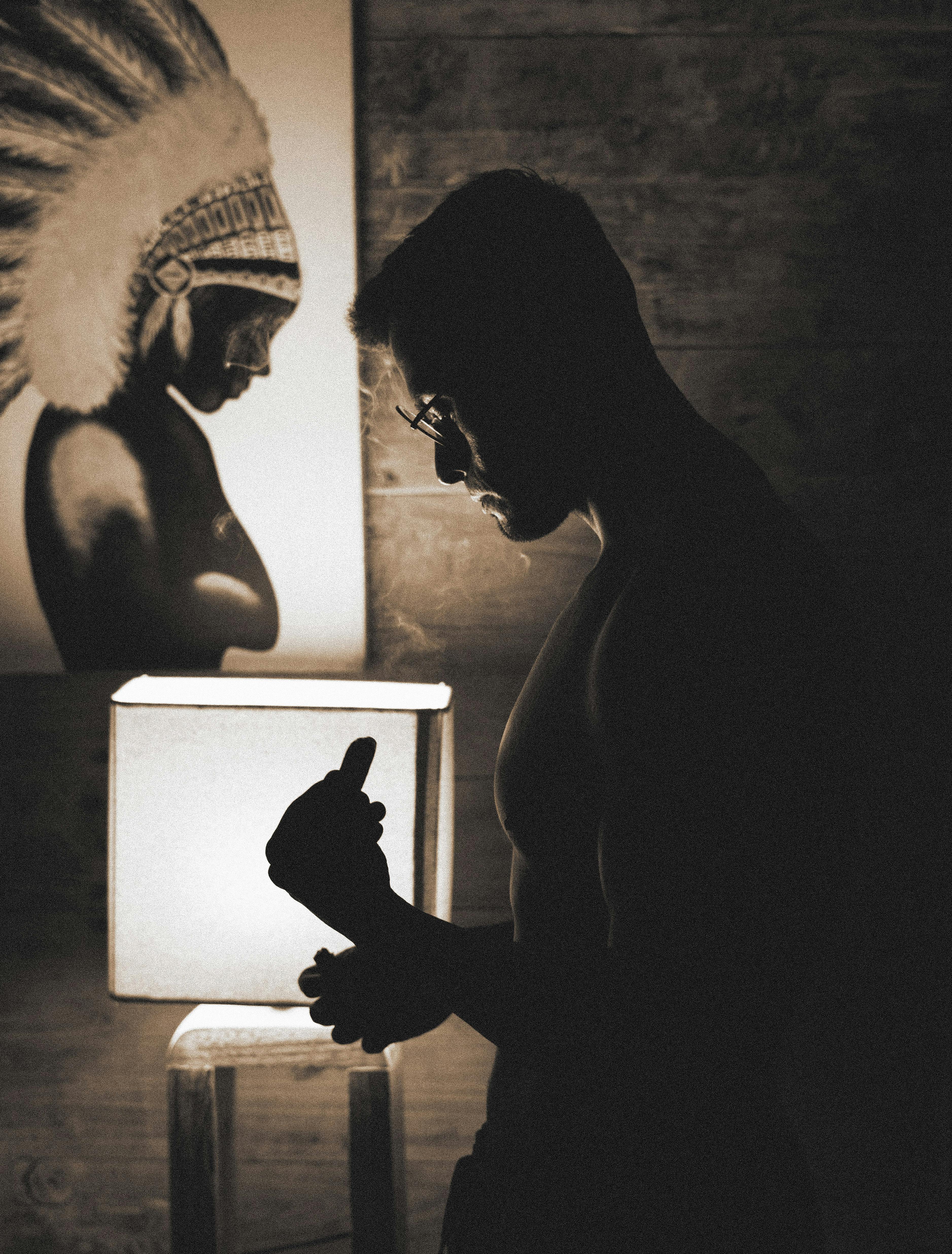 A shirtless man in silhouette stands beside a lit lamp and artwork in a dimly lit room.