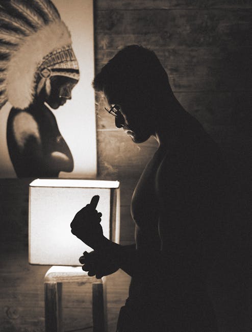 A shirtless man in silhouette stands beside a lit lamp and artwork in a dimly lit room.
