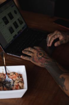 Tattooed individual working on a laptop with a breakfast bowl nearby, capturing a cozy morning vibe.