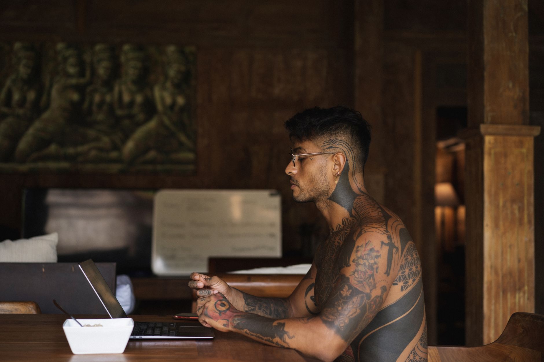 Tattooed man with eyeglasses sitting at a desk using a laptop in a dimly lit room.