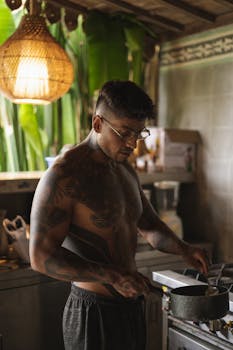 A shirtless man with tattoos cooking and stirring a pot in a rustic indoor kitchen.