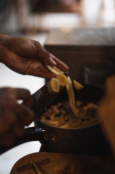 A person slices bananas over a pot in a cozy kitchen setting, creating a warm culinary scene.