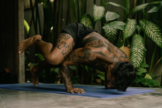 A tattooed man performs a yoga pose on a mat surrounded by lush green plants, showcasing strength and balance.