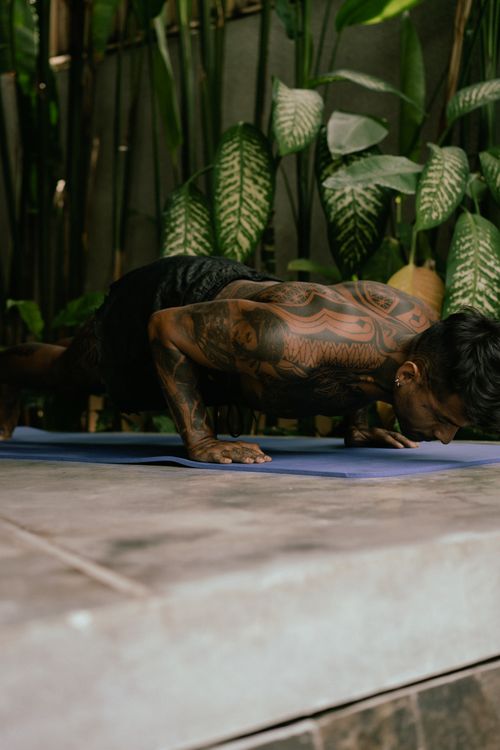 Free A tattooed man performs push-ups on a mat outdoors surrounded by lush plants. Stock Photo