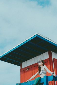 Woman in sunglasses relaxing at a colorful lifeguard station under a clear sky.