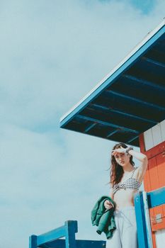 Young woman in a bikini and sweatpants standing by a colorful beach lifeguard tower on a sunny day.